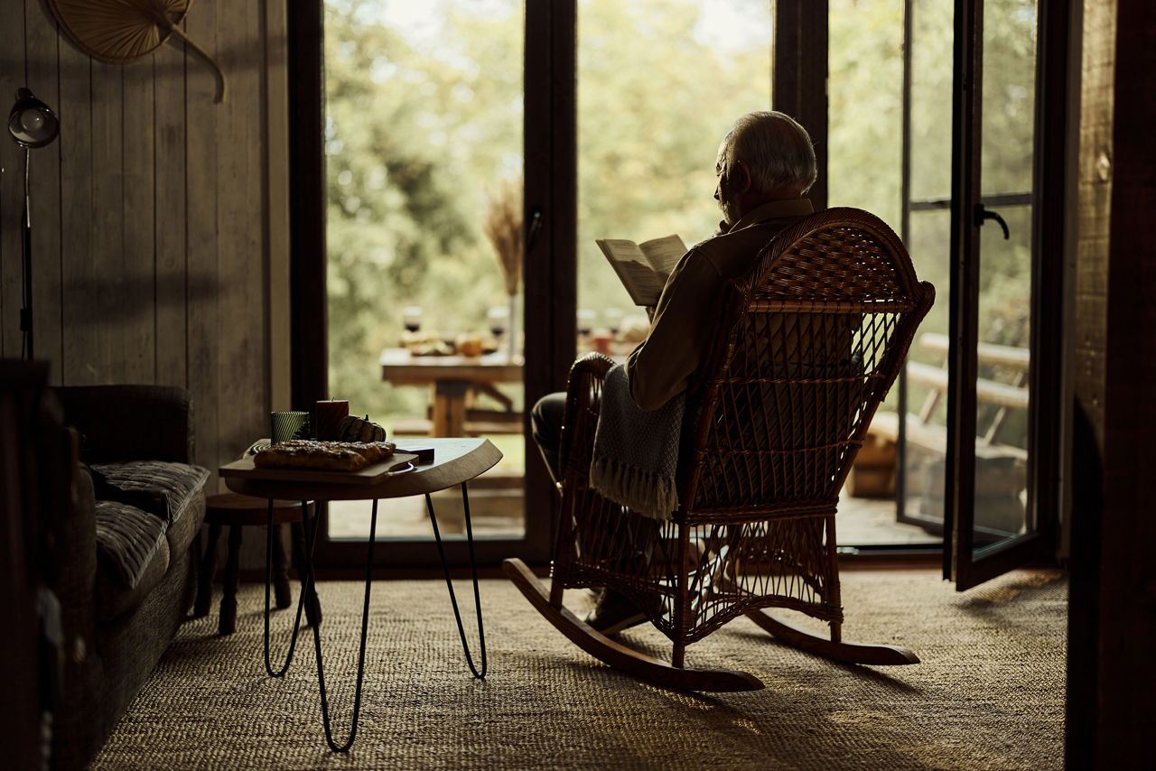 Rear view of mature man reading a novel in rocking chair by the window in the living room.